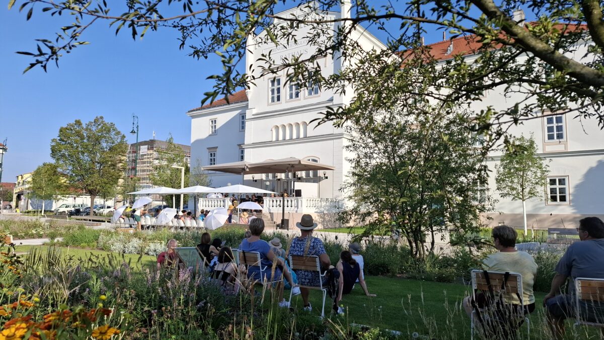 Menschen sitzen unter einem Baum und schauen auf ein Konzert auf einer Terrasse.